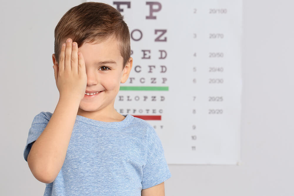 Junge hält sich beim Sehtest ein Auge zu und lächelt, im Hintergrund eine Sehprobentafel