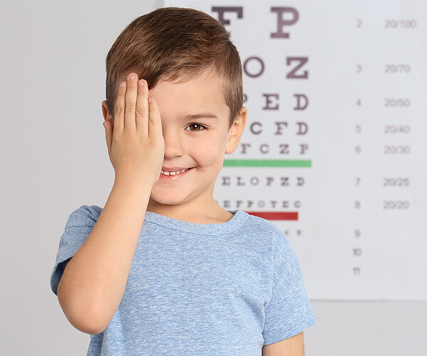Junge hält sich beim Sehtest ein Auge zu und lächelt, im Hintergrund eine Sehprobentafel
