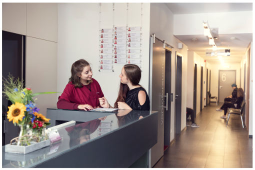 Zwei Frauen im Gespräch an einem Empfangstresen in einer Arztpraxis, im Hintergrund eine Personen